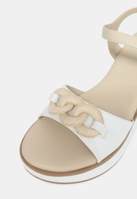 Sandali bianchi con suola beige, caratterizzati da un dettaglio di cinturino intrecciato e una singola fibbia. Texture liscia e design minimalista.
