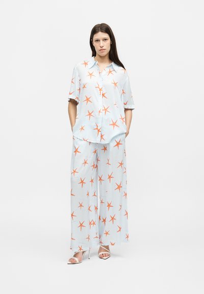 Femme portant une chemise bleu clair assortie et un pantalon à jambes larges avec des motifs d'étoiles de mer orange, debout les mains dans les poches et portant des talons blancs.
