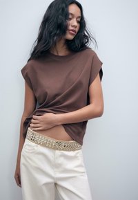 Vrouw met lang golvend zwart haar, gekleed in een bruin mouwloos topje en witte broek met een decoratieve gouden en spiegelachtige riem, poserend tegen een lichte muur.