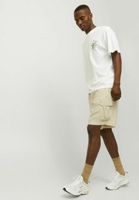 T-shirt blanc à manches courtes avec un imprimé graphique, associé à un short cargo beige avec des poches latérales, et des baskets blanches avec des chaussettes beiges.