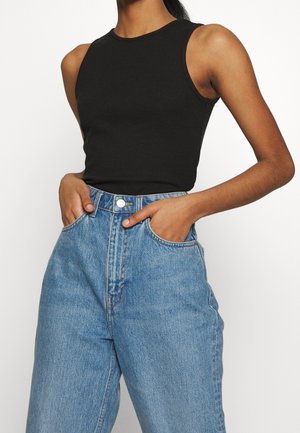 Woman wearing sleeveless black ribbed top tucked into high-waisted loose-fitting blue jeans with hands in front pockets.
