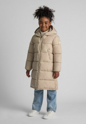 Niña joven con cabello rizado que lleva un abrigo acolchado beige, jeans claros, zapatillas blancas, sonriendo frente a un fondo gris liso.
