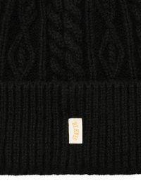 Zwarte gebreide beanie met een gestructureerd kabelontwerp. Beschikt over een geribde manchet en een klein, opgenaaid label met "BLKWS" in gouden letters.