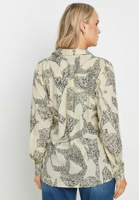 Cream blouse with a black abstract pattern, long sleeves, buttoned cuffs, and a back yoke detail. Smooth fabric with a relaxed fit.