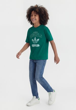 Sretno dijete s kovrčavom kosom hoda, nosi zelenu majicu Adidas Originals, plave traperice i bijele tenisice sa zelenim detaljima.