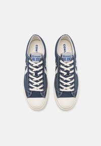 Námořnické plátěné tenisky s bílým gumovým špičákem, krémovými tkaničkami a bočními pruhy. Text „Converse All Star“ na jazyku a vnitřní vložce.