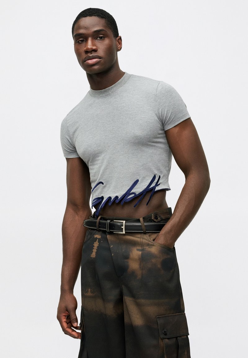 Grijze, kortgebouwde t-shirt met een ronde halslijn, voorzien van blauw geborduurde tekst. G gedragen met donkere, gemêleerde cargobroeken en een zwarte riem.