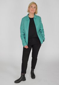 Teal Lederjacke mit Reißverschluss, schwarzes Oberteil, schwarze Hose und schwarze Stiefeletten. Glatte Textur, taillierte Passform und minimales Design.