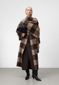 Cappotto di lana scozzese marrone e beige con una vestibilità over, abbinato a una gonna marrone scuro e stivali neri con tacco. Tiene in mano una borsa nera.