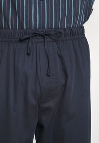 Pantaloni blu navy con vita elasticizzata e cordoncino, caratterizzati da una texture liscia e tasche laterali. La camicia ha righe verticali di colore teal.