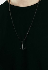 Collana con pendente ad ancora nera su una sottile catena d'argento attorcigliata, indossata sopra una t-shirt nera. Il pendente ha una finitura lucida.
