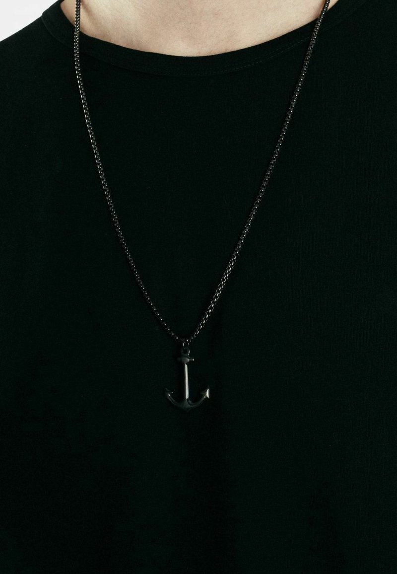 Collana con pendente ad ancora nera su una sottile catena d'argento attorcigliata, indossata sopra una t-shirt nera. Il pendente ha una finitura lucida.