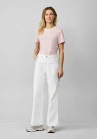 Jasnoróżowa, w paski koszulka z napisem "happy" wykonanym kursywą, w zestawieniu z białymi, szerokimi spodniami z wysokim stanem. Modelka stoi z rękami w kieszeniach.