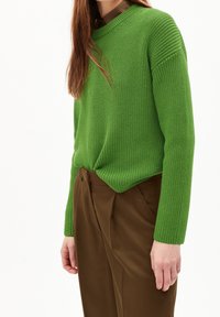 Frau in grünem, langärmligem Strickpullover, der in hochtaillierte braune Hosen gesteckt ist, steht vor einem schlichten weißen Hintergrund.