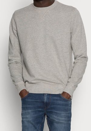 Mann trägt hellgrauen Sweatshirt mit Rundhalsausschnitt und blaue Jeans mit den Händen in den Taschen vor einfachem Hintergrund.