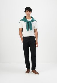 Junger Mann mit dunklem lockigem Haar, der ein weißes Logo-T-Shirt, schwarze Hose, braune Sneaker und einen um die Schultern gebundenen grünen Pullover trägt und steht.