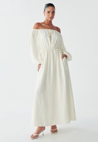 Una donna sta in piedi con sicurezza, indossando un vestito bianco fluido con spalle scoperte e vita elastica, abbinato a tacchi bianchi con strappi.