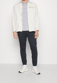 Vaaleanbeige puuvillatakk, raidallinen harmaa poolopaita, tummat housut joissa taskut, ja valkoiset lenkkarit keltaisilla yksityiskohdilla. Yksinkertainen, rento asu.