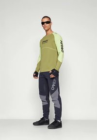 Grünes und hellgrünes langärmeliges Hemd mit schwarzem Logo, kombiniert mit dunkelblauen Hosen mit grauen Akzenten und schwarzen Schuhen. Schwarze Sonnenbrille.