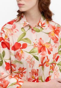Floral blouse with a pastel pink background, featuring vibrant red and green floral patterns. Buttoned collar, lightweight fabric, long sleeves.