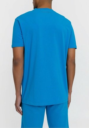 Homme portant une chemise bleu vif à manches courtes et un pantalon bleu assorti, vu de dos sur un fond clair uni.