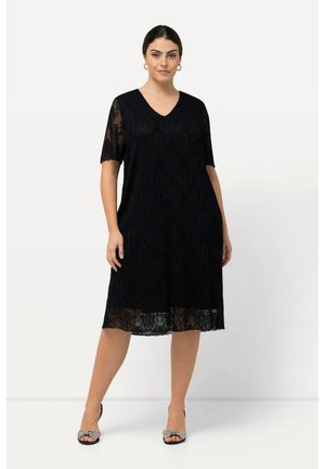 Donna che indossa un vestito di pizzo nero a metà coscia con maniche corte e scollatura a V, abbinato a tacchi aperti neri decorati con strass.
