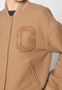 Brązowa kurtka z żeberkowym wzorem, z teksturowanym logo "G" na piersi, z zatrzaskami i bocznymi kieszeniami. Miękki materiał z dopasowanym krojem.