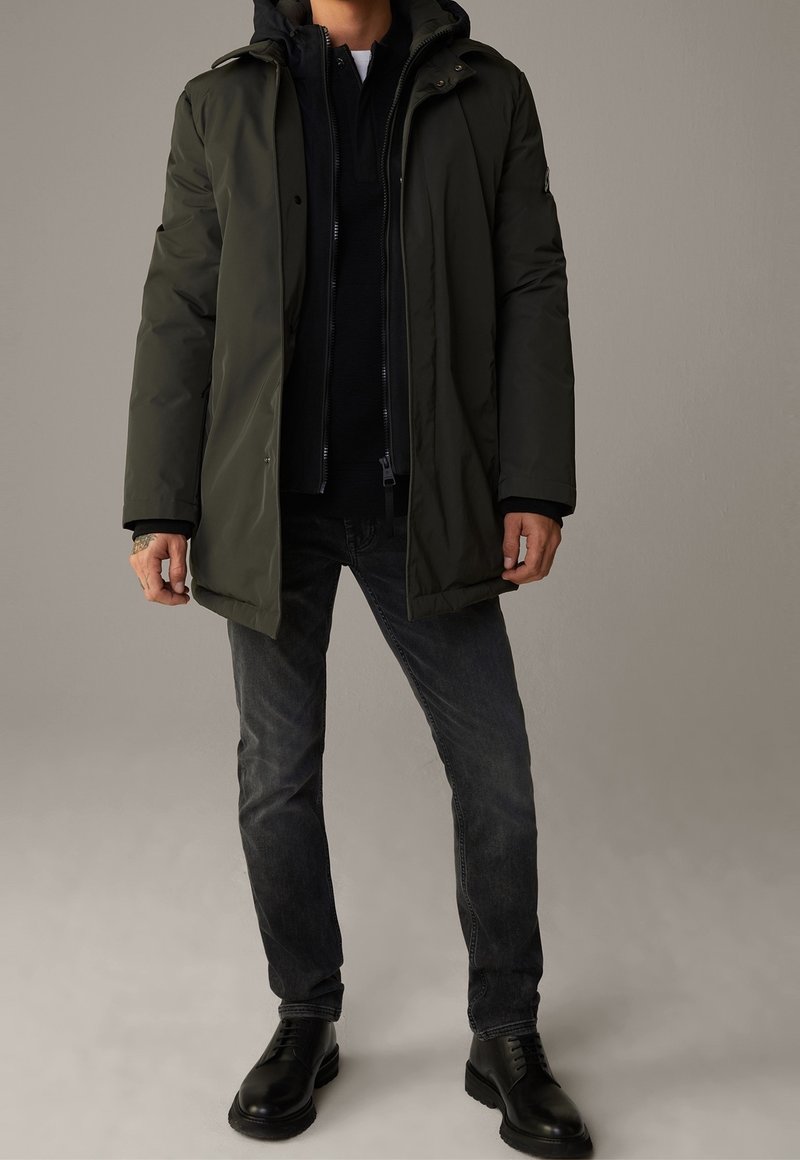 Homme portant une veste d'hiver à capuche vert foncé, un pull zippé noir, un jean noir délavé et des bottes noires à lacets, debout devant un fond uni.