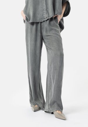 Person wearing loose gray washed denim pants and matching button-up shirt with hands in pockets, light gray pointed shoes visible.