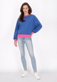 Felpa blu con polsini e orlo rosa, presenta una vestibilità comoda e maniche lunghe, abbinata a jeans skinny azzurri chiari e sneakers bianche.