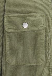 Tessuto a coste in verde oliva con righe verticali, dotato di una grande tasca a toppa e un dettaglio con bottone a pressione argento.