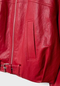 Gros plan sur une veste en cuir rouge montrant une fermeture éclair argentée, une poche latérale et une ceinture réglable avec boucle métallique en bas.