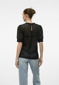 Top de encaje negro con mangas cortas abullonadas y detalle de abertura en la espalda, combinado con pantalones vaqueros de denim azul claro. El encaje tiene un patrón ondulado.