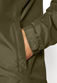 Veste imperméable olive avec une poche et des poignets élastiqués. Texture lisse avec un éclat subtil et des détails de couture renforcés.