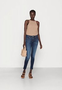 Canotta marrone a coste, jeans aderenti blu e sandali marroni. Tiene una piccola borsa intrecciata. Design semplice con un aspetto casual.