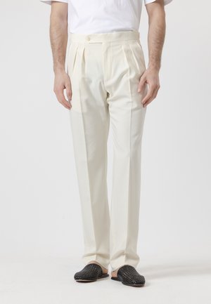 Hombre con pantalones de talle alto, plisados y de color blanco roto, y zapatos negros tejidos sin cordones, frente a un fondo liso.