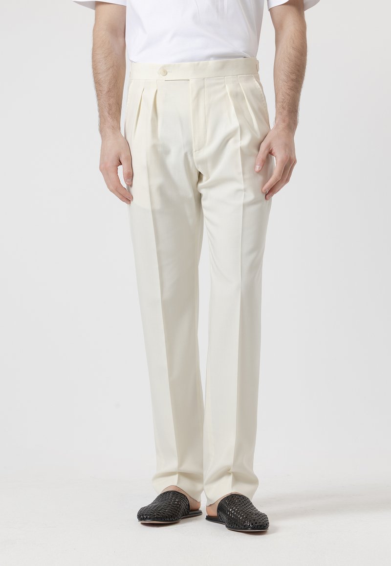 Hombre con pantalones de talle alto, plisados y de color blanco roto, y zapatos negros tejidos sin cordones, frente a un fondo liso.