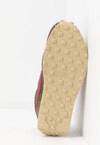 Semelle d'une basket avec une semelle en caoutchouc beige comportant des clous carrés et des numéros de brevet, chaussure inclinée contre un fond d'angle blanc.