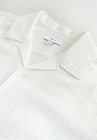 White short-sleeve shirt with a textured grid pattern, button closure, and a pointed collar. Brand label "NEXT" inside the collar.