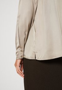 Beige Satinbluse mit langen Ärmeln, Knopfarsch, seitlichen Schlitzen und einer glatten Textur, kombiniert mit einem gerippten dunkelbraunen Rock.