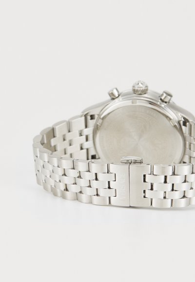 Silverfärgad klocka i rostfritt stål med en länkad armbandsdesign, polerad yta, rund boett och en synlig krona med en ädelstensdetalj.