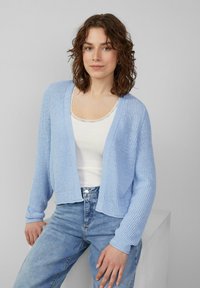 Lichtblauwe gebreide cardigan met een losse pasvorm, open front en ribbelstructuur. Gepaard met een witte tanktop en lichtblauwe spijkerbroek.