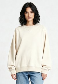Sweat-shirt ample beige clair avec manches raglan, poignets et ourlet côtelés, texture douce et détails de couture minimalistes. Associé à un jean bleu.