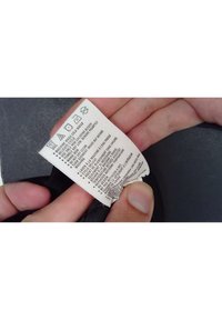 Štítek na textilu, bílý podklad, černý text uvádějící pokyny pro údržbu se symboly pro praní, sušení a žehlení. Ruka držící štítek.