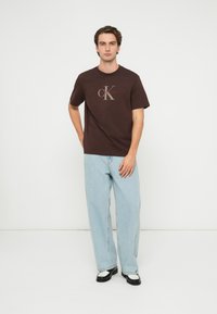 T-shirt de algodão castanha com um logótipo da Calvin Klein cinza claro no peito, combinado com jeans azul claro e sapatos pretos com detalhes brancos.