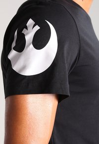 Černé tričko s krátkým rukávem na osobě s velkým bílým symbole Rebel Alliance na horním rukávu.