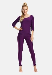 Frau steht barfuß da, trägt ein eng anliegendes lila Langarmshirt und passende Leggings, mit der Hand an der Hüfte vor einem einfarbigen Hintergrund.