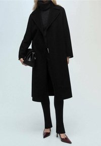 Long manteau noir avec un large col, porté sur un col roulé foncé, associé à un pantalon noir sur mesure et des escarpins bordeaux à bout pointu.