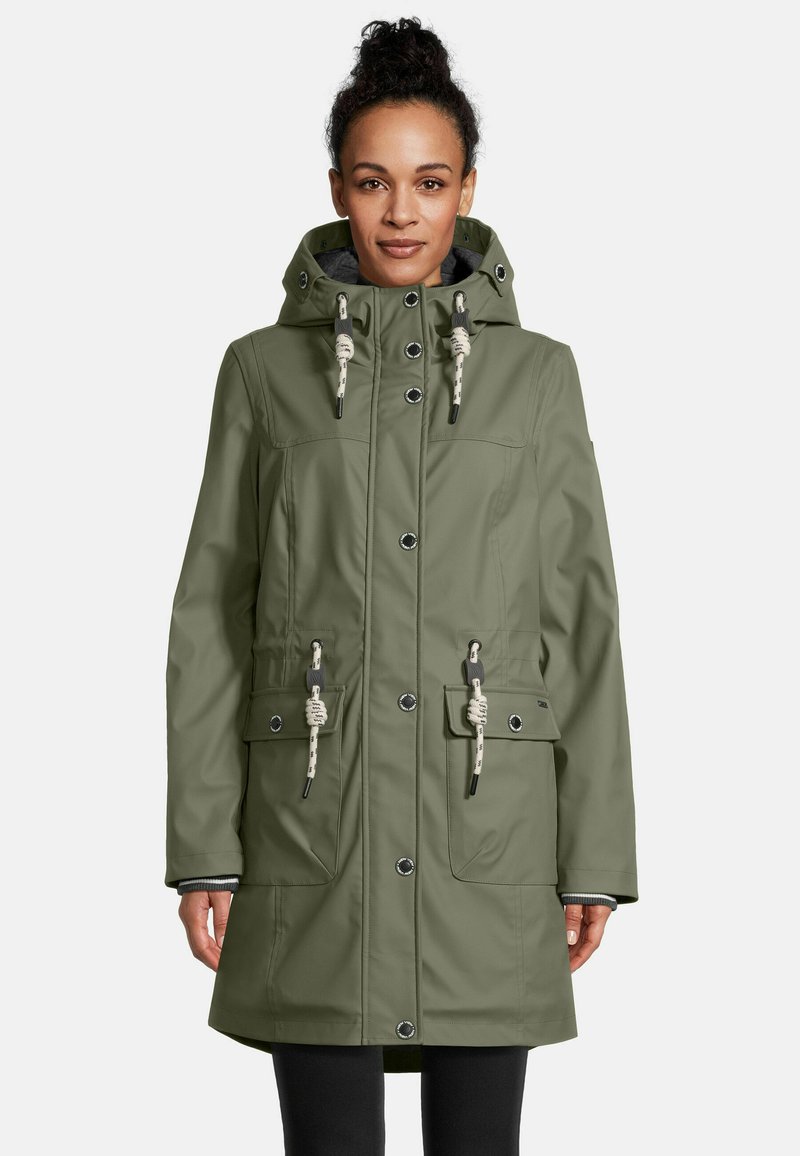 Een vrouw in een olijfgroene regenjas met capuchon, voorzien van trekkoorden en drukknoopjes, staand voor een eenduidige lichte achtergrond.