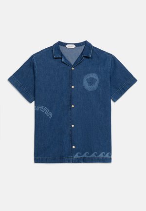 Chemise en denim bleu foncé à manches courtes avec boutonnière sur le devant, col, logo Méduse sur la poitrine, et motifs ondulés et géométriques près de l'ourlet et sur les côtés.